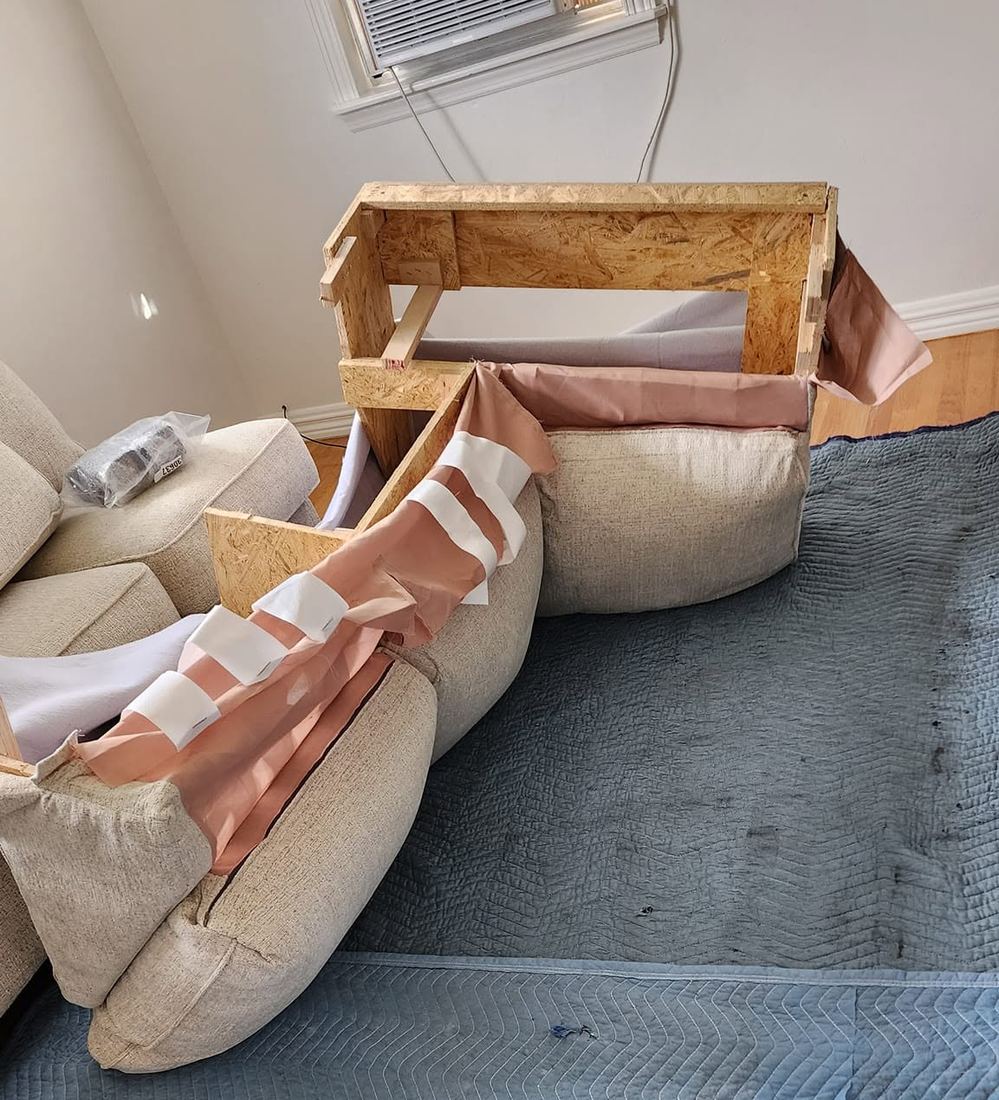 Beige sofa section with its wood frame exposed and pink lining folded back with white tape, on grey blankets