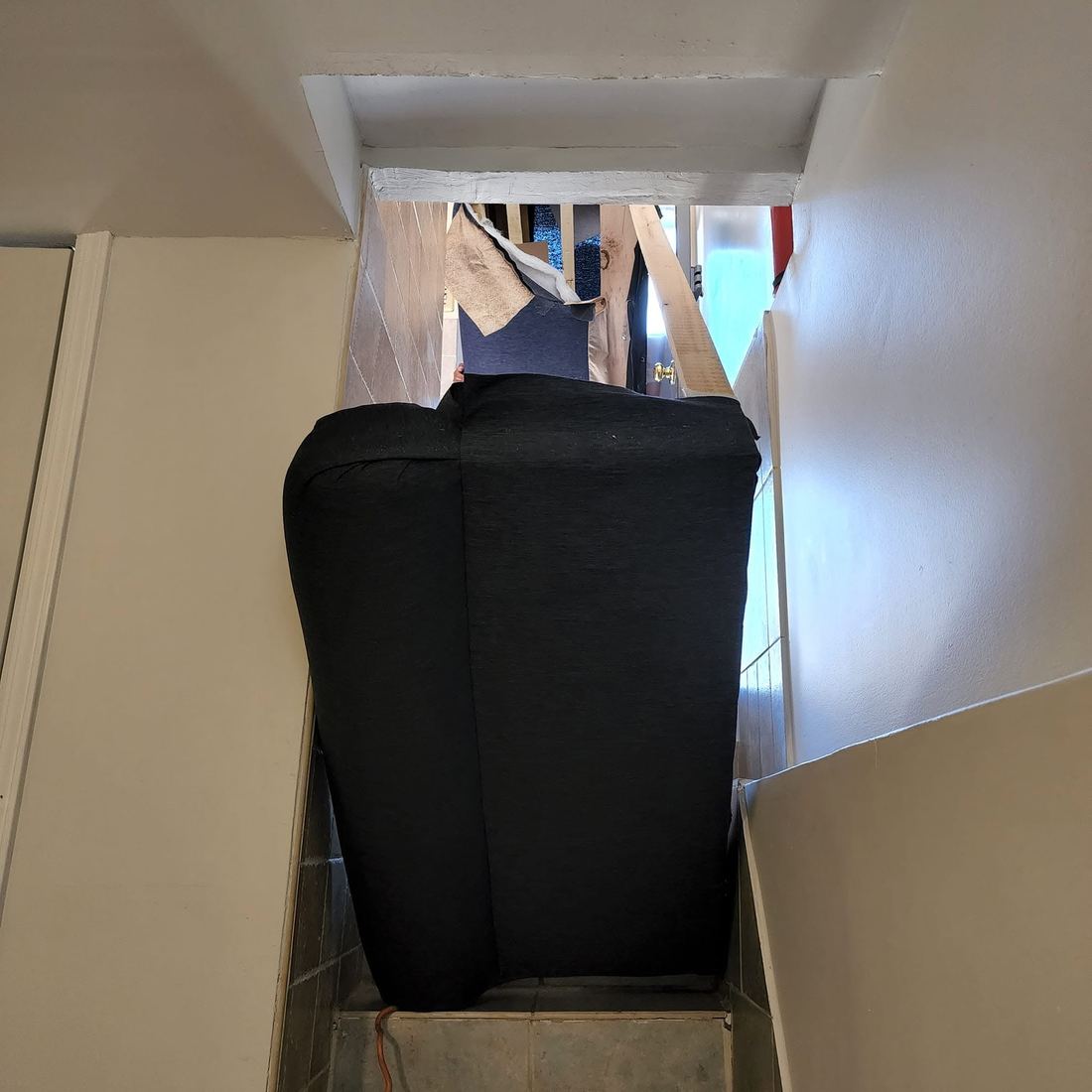 Looking up a narrow tiled stairway at a black sofa section standing upright under a low ceiling beam