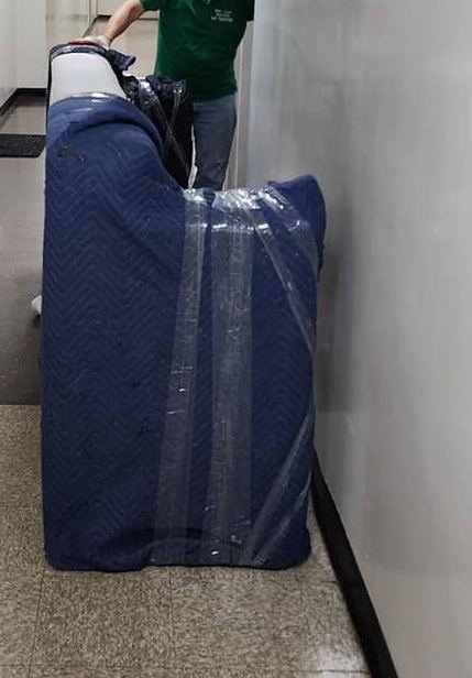 Sofa wrapped in blue padded blankets and plastic, stood on end in a narrow hallway with a crew member