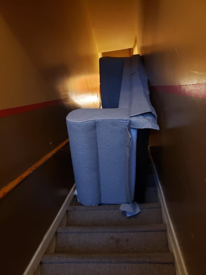 Light blue upholstered sofa section standing on end on the steps of a narrow, dimly lit carpeted staircase