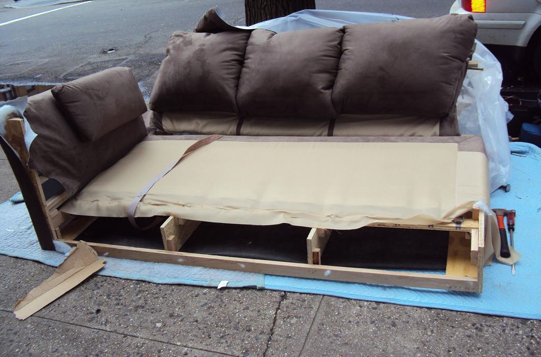 Brown microfiber sofa on blue blankets on a city sidewalk, seat deck and wood base frame exposed, hammer beside it