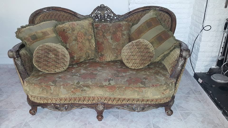 Ornate carved wood-frame sofa with floral tapestry upholstery and fringed square and round pillows on a tiled floor