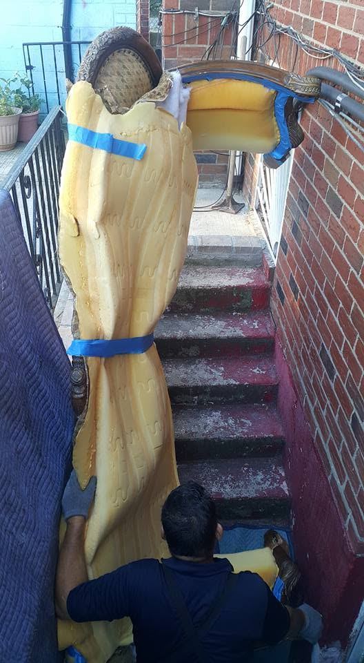 Carved wood sofa wrapped in yellow foam and blue tape, held on end on outdoor steps by a worker seen from behind