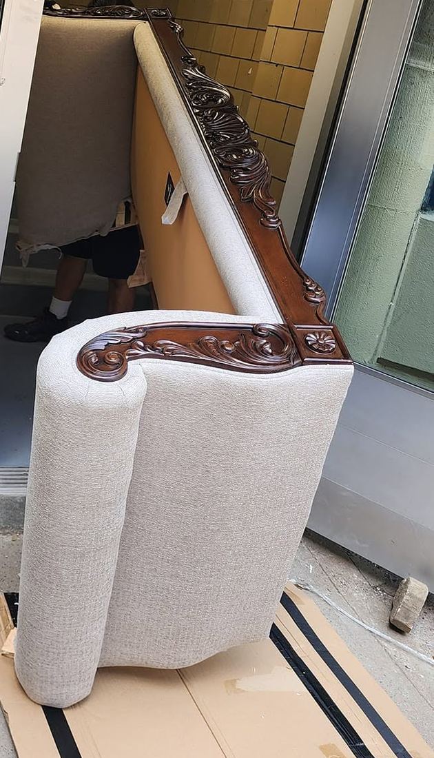 Beige sofa with carved dark wood trim tipped vertically on cardboard at an entry door, crew member behind it