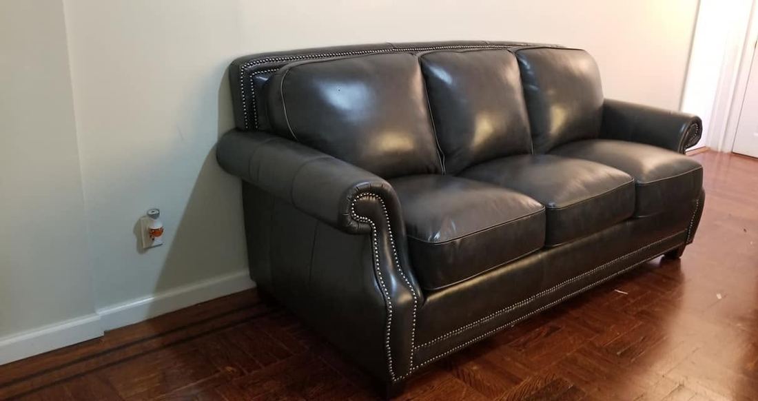 Charcoal leather three-seat sofa with rolled arms and nailhead trim against a white wall on a parquet floor
