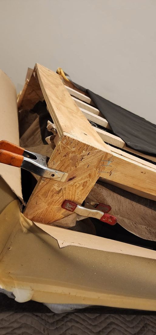 Close-up of orange and red spring clamps gripping a sofa's plywood frame joint beside foam padding
