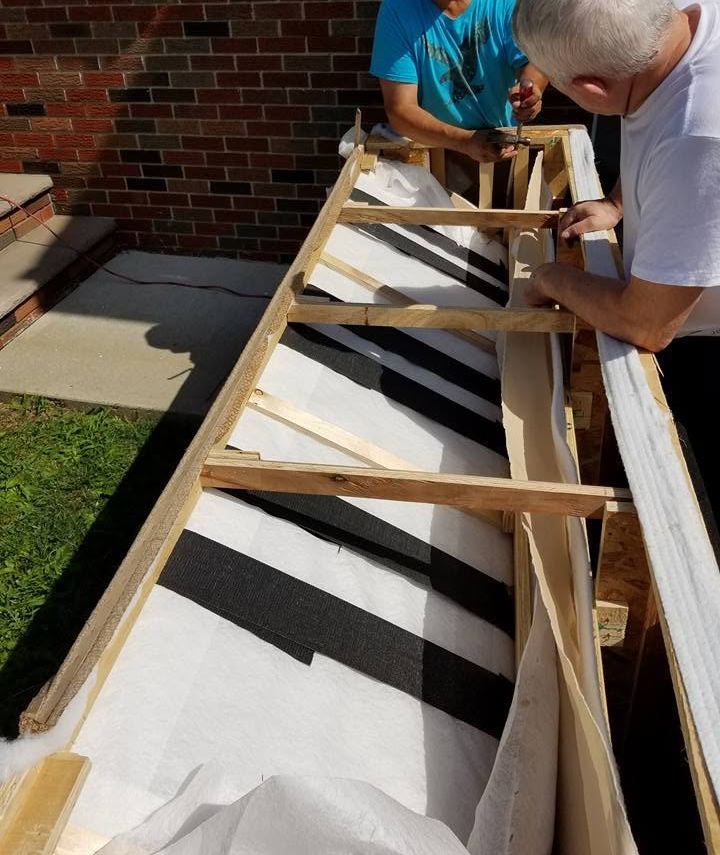 Two craftsmen lean over an open wooden sofa frame lined with webbing and batting outdoors