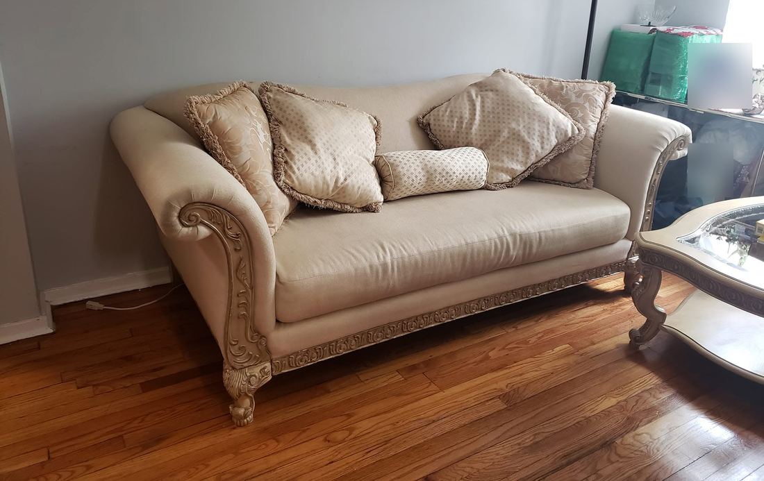 Cream upholstered sofa with carved wooden arms and base and fringed pillows, set on a hardwood floor