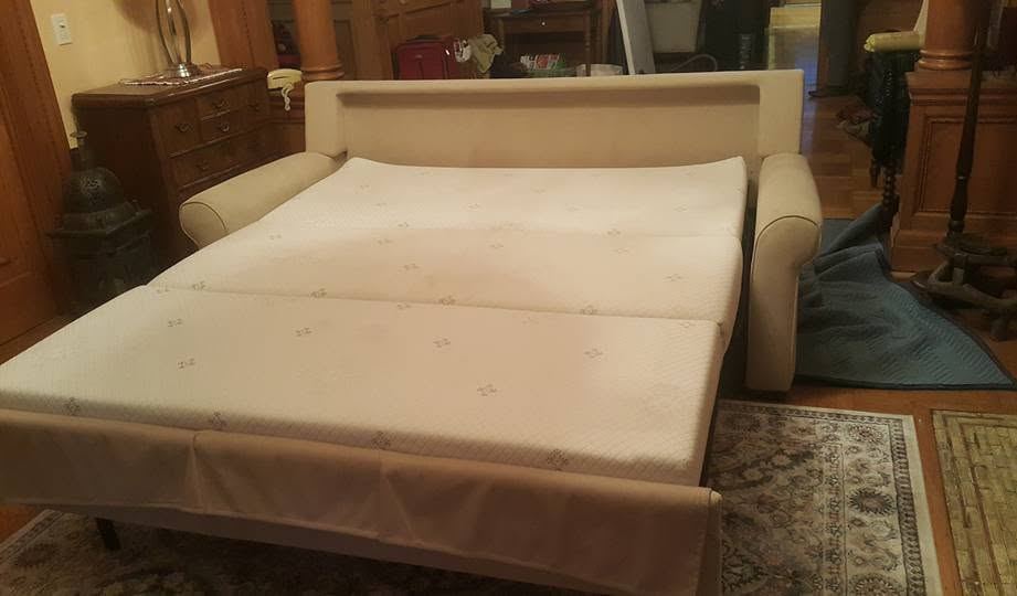 Cream sleeper sofa pulled out into a bed, patterned mattress laid flat over a rug, a quilted pad on the floor beside it