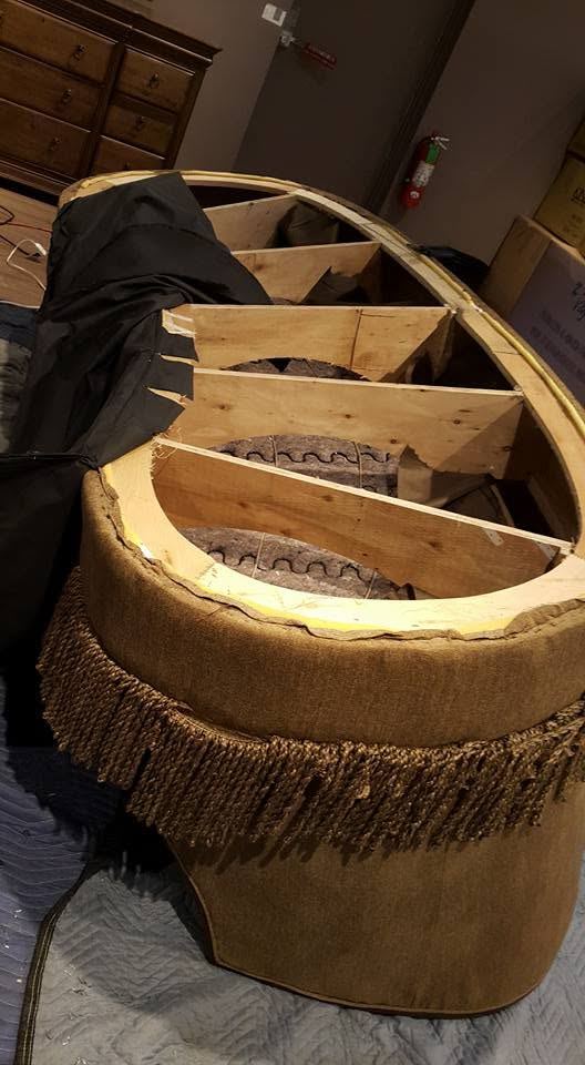 Curved sofa with a brown fringe skirt, upholstery peeled back to show curved wooden ribs and springs, on padded blankets