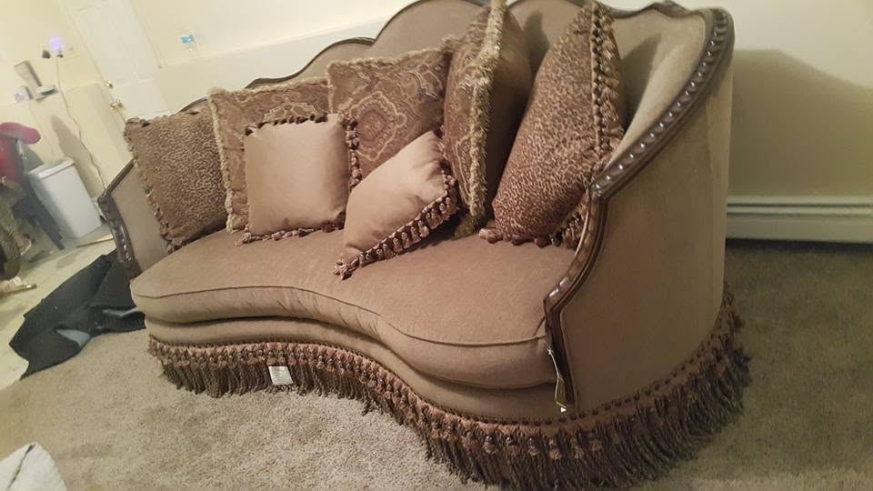 Tan curved-back sofa with carved wood trim, a brown fringe skirt and fringed accent pillows on beige carpet