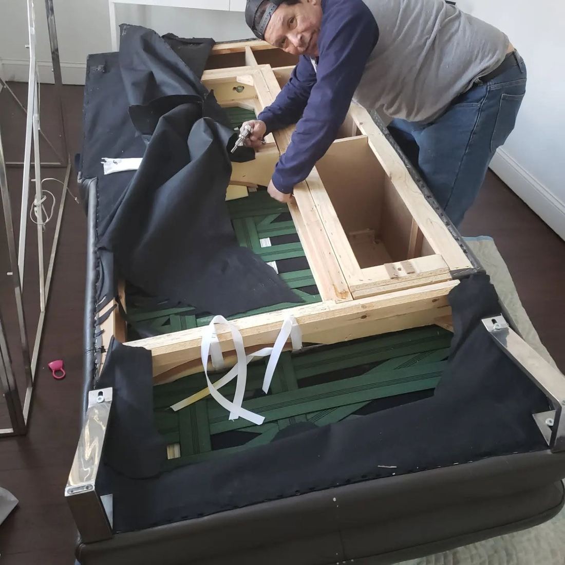 Doc Juan in a backwards cap works on the exposed wood frame and green webbing of an upturned leather sofa