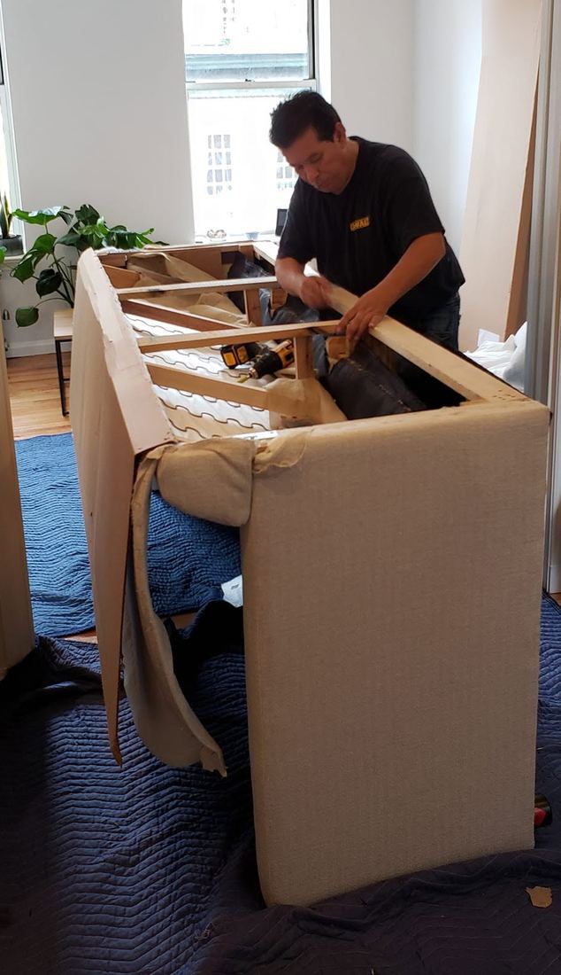 Doc Juan works on the opened wood frame of a light gray sofa set on blue protective blankets