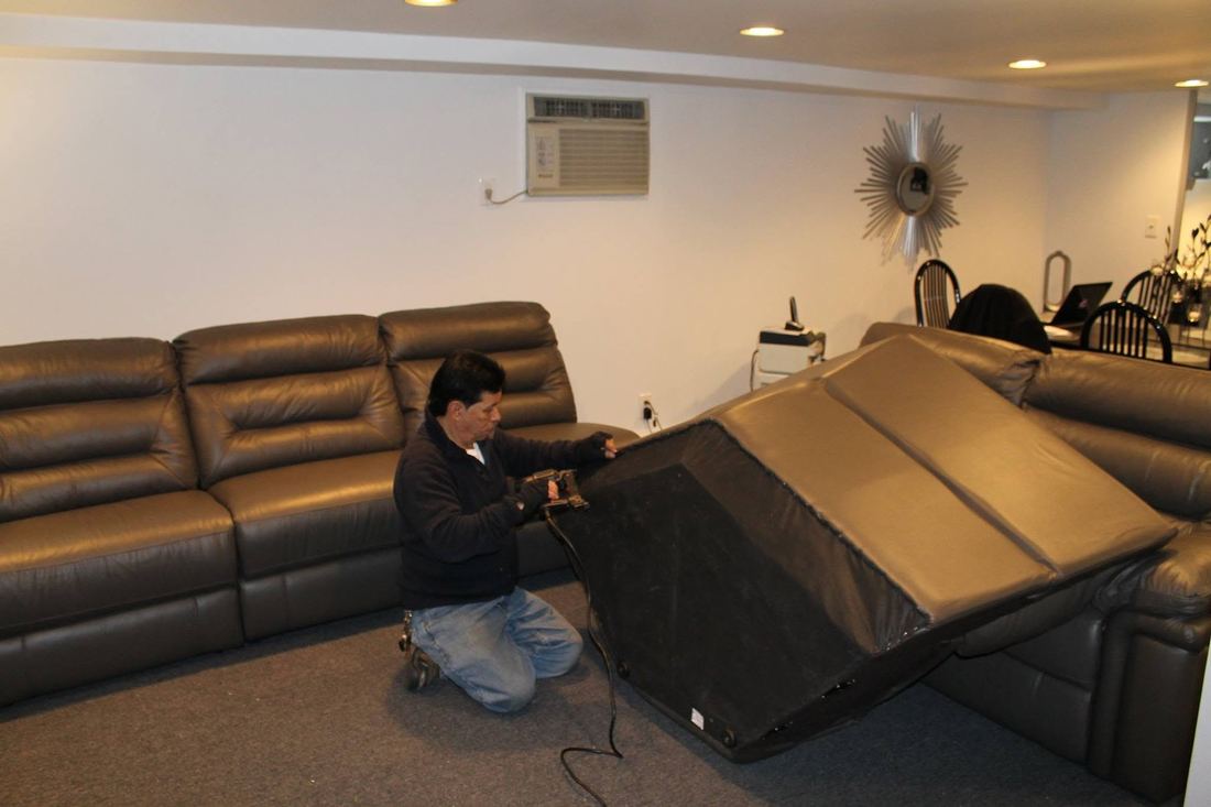 Doc Juan kneels beside a gray leather sectional, working a corded tool along the edge of one tipped-back section