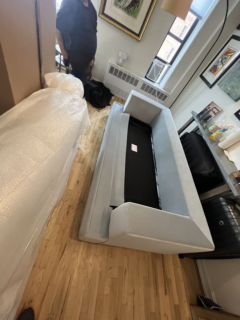 Light gray sleeper sofa with cushions removed and black sleeper deck exposed, on a hardwood floor in a sunny apartment