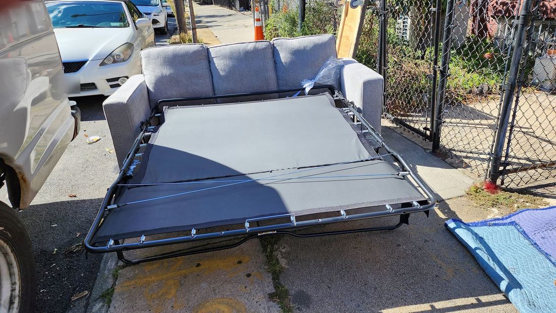 Gray three-seat sleeper sofa outdoors on a sidewalk with its metal fold-out bed frame fully extended