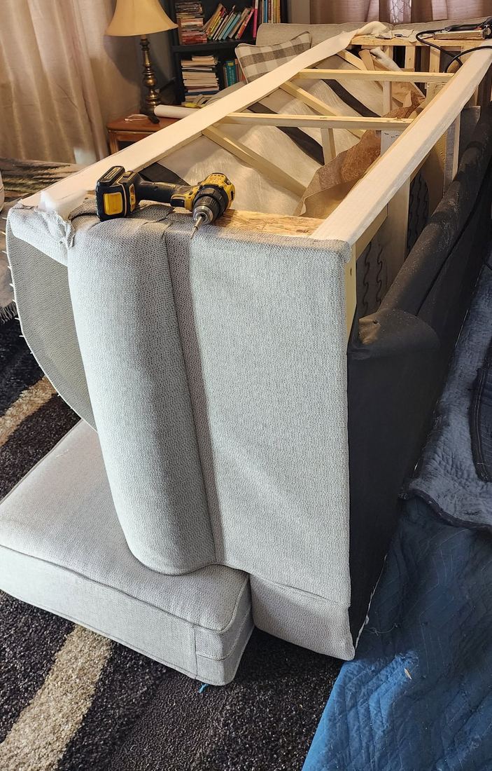 Grey fabric sofa with the back upholstery opened, showing the wood frame, with a cordless drill on the arm