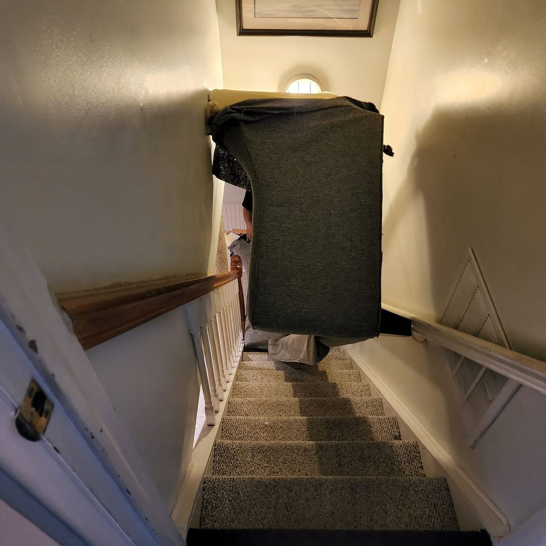 Looking down a narrow carpeted staircase at a dark grey sofa section carried between the walls