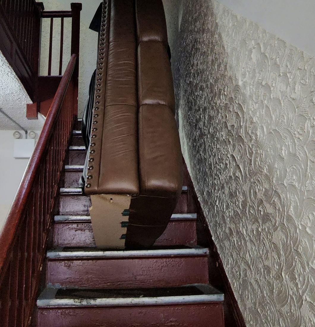 Brown leather sofa with nailhead trim held upright on a narrow staircase with a red wooden banister