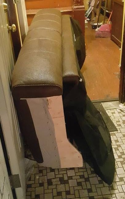 Brown leather sofa section with an exposed wood frame end, stood upright on a tiled hallway floor by an open door