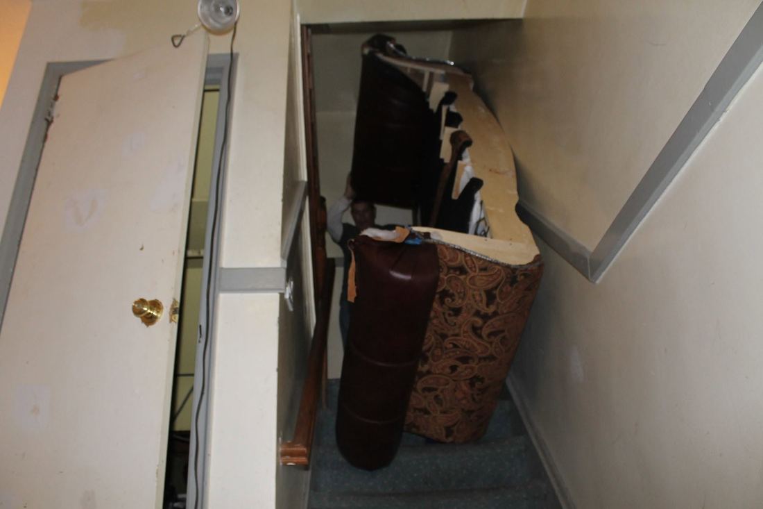 A crew member lifts a carved sofa section with paisley fabric and dark leather through a narrow carpeted stairwell