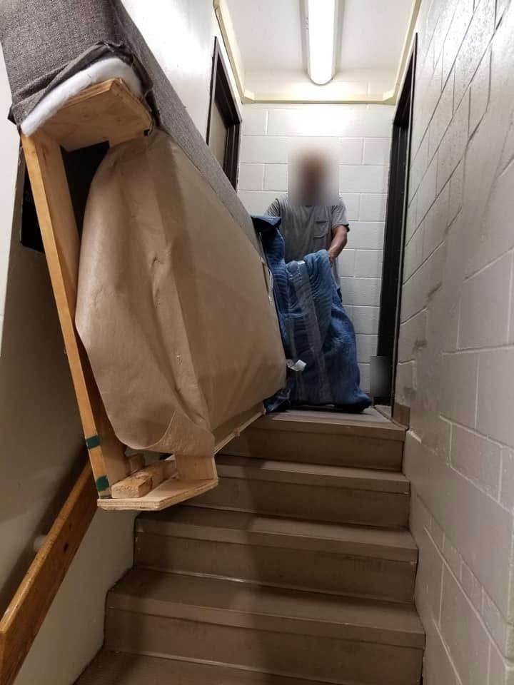 Sofa wrapped in brown paper tipped on its end on the stairs of a narrow cinder-block stairwell, a worker behind it
