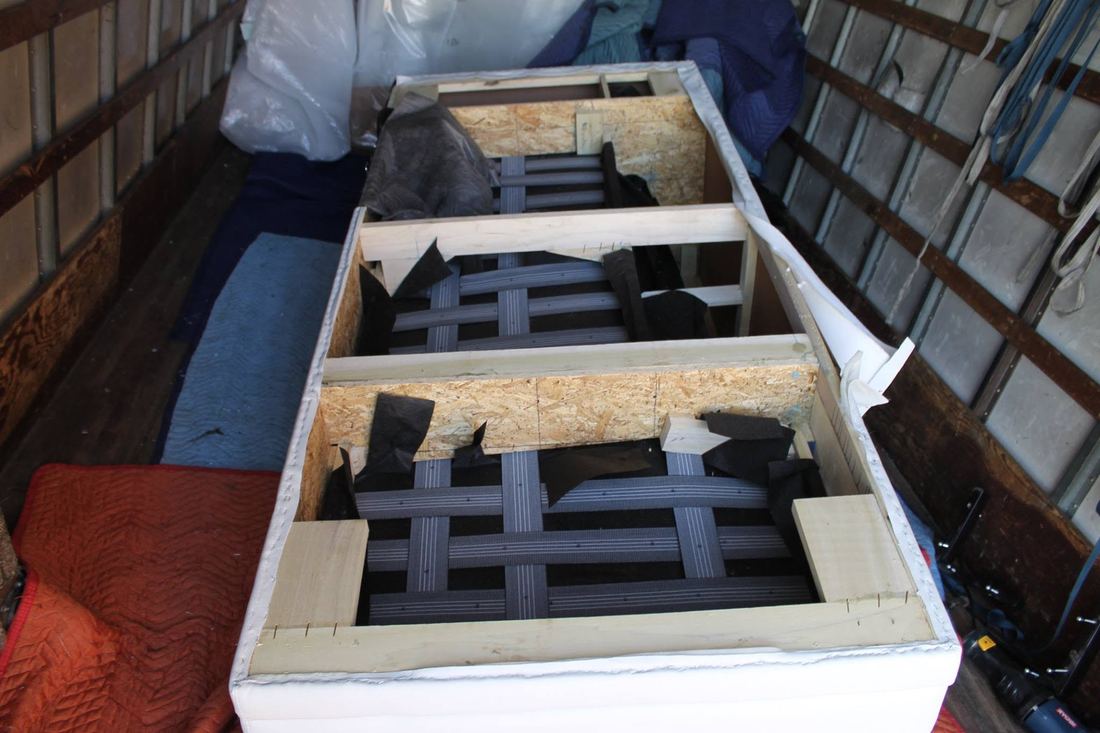 Inside a box truck, white sectional base frames with undersides opened, showing gray webbing, wood rails and plywood