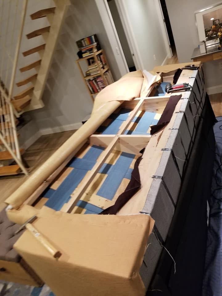 Long sofa with its tan fabric folded back over a wooden frame and blue webbing, a power tool resting on the rail
