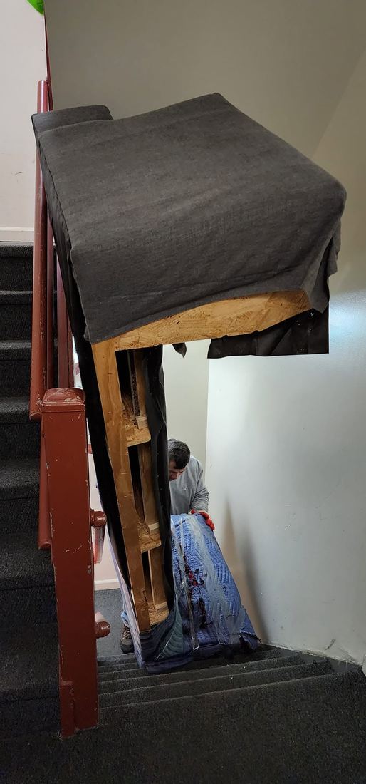 Sofa frame with its dust cover showing, upended in a narrow carpeted stairwell beside a red railing