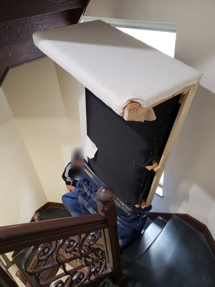 Cream sofa section stood on end and wrapped in blue padded blankets, angled through a curved stairwell