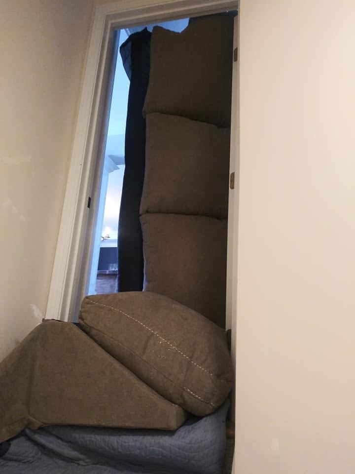 A taupe sofa stands upright on its end inside a narrow doorway, a cushion and a detached piece resting on a quilted pad