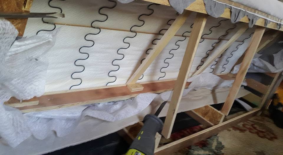 Underside of an opened sofa showing zigzag springs and wood braces, with a green reciprocating saw at the frame