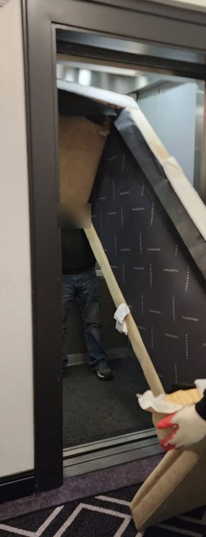 Sofa section with its black dust cover showing, tilted diagonally into an elevator by a worker's gloved hand