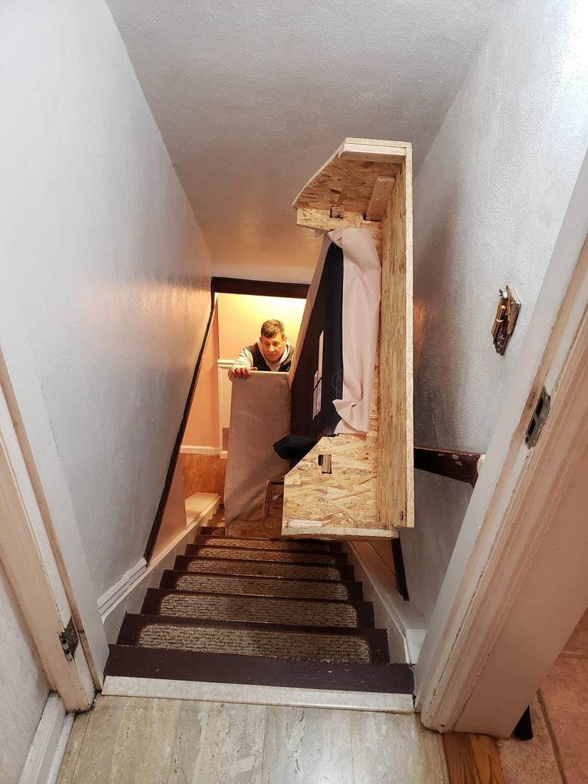 Sofa frame with its upholstery partly removed angled through a narrow carpeted staircase as a crew member steadies a panel