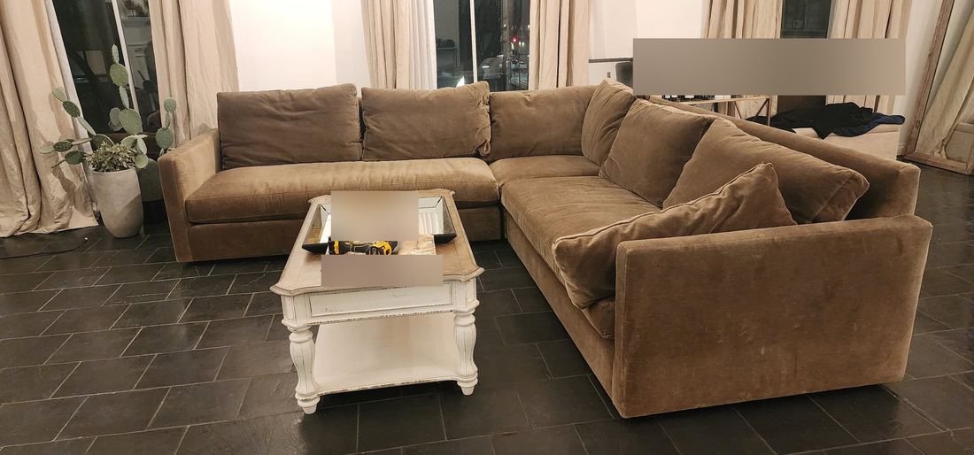 Taupe velvet L-shaped sectional around a distressed white coffee table on a dark slate tile floor