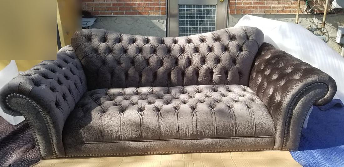 Button-tufted gray Chesterfield-style sofa with rolled arms and nailhead trim on quilted pads beside a brick wall
