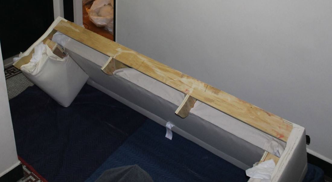 Long white sofa section with its wood frame exposed, angled across a small apartment-building hallway on padded blankets