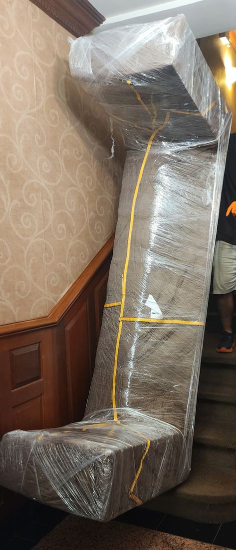 Sofa wrapped in plastic and padding, stood on end at the base of a carpeted staircase with patterned wallpaper