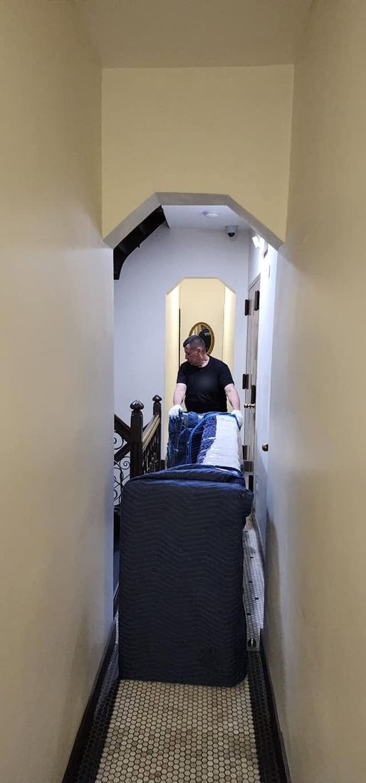 Sofa wrapped in blue padded blankets stands on end in a narrow hallway while a gloved crew member steadies it