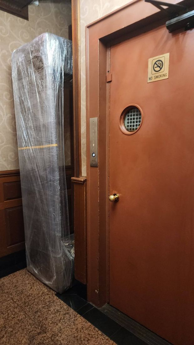 Dark sofa wrapped in clear plastic, stood upright on its end next to a brown elevator door