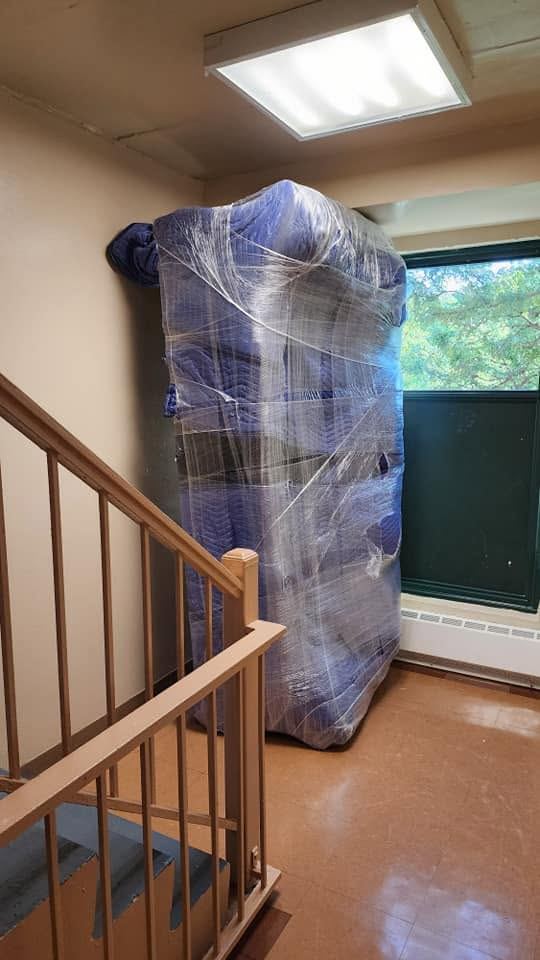 Sofa wrapped in blue padded blankets and plastic film stands on end beside a stairwell railing